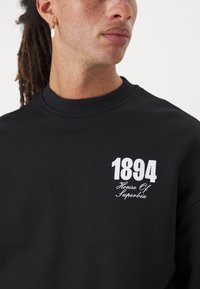Sudor neopren negru, cu guler rotund, manșete și tiv din tricot, având un text grafic alb "1894" și "House Of Superbia". Textură netedă.
