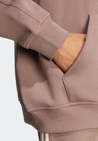 Dammig rosa sweatshirt med ribbade manschetter, en kenguruficka och strukturerad tyg. Nära vy visar handen som vilar i fickan.