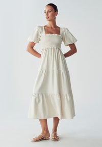 Moteris stovi su rankomis ant klubų, vilkėdama smėlio spalvos midi suknelę su pūstu rankovėmis ir šlepetėmis prie vienspalvio šviesaus fono.