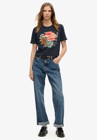 T-shirt graphique bleu marine avec un design de tigre vintage, associé à un jean taille haute bleu et des chaussures noires. Manches retroussées sur le jean.
