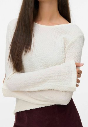 Femme aux longs cheveux foncés portant un haut blanc texturé à manches longues avec de larges poignets et un pantalon bordeaux foncé, bras croisés.