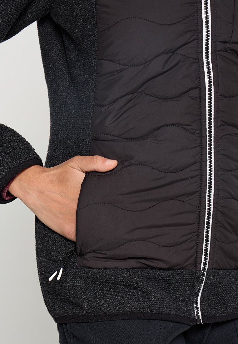 Mano parzialmente dentro della tasca di una giacca nera trapuntata con una cerniera frontale visibile e maniche in tessuto texture.