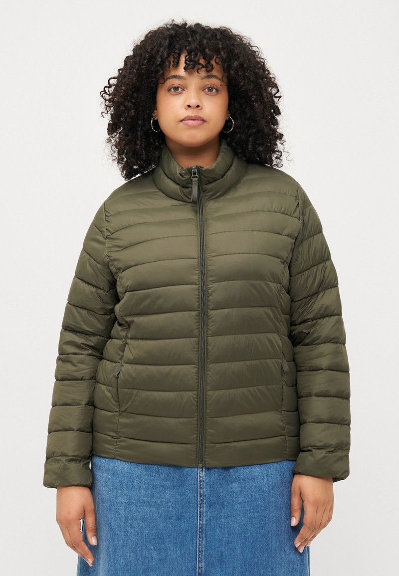 Olijfgroene pufferjack met een hoge kraag, horizontale stiksels en ritsluiting. Draag over een blauwe denim rok.