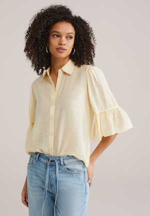 Femeie cu păr creț, purtând o bluză crem cu nasturi și mâneci cu volane și blugi albastru deschis, stând în fața unui fundal simplu.