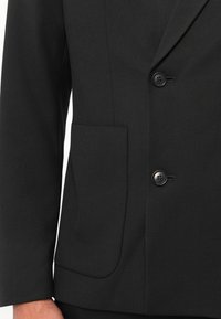 Chaqueta de traje negra con un diseño de un solo botón, dos botones y un bolsillo lateral. La tela es suave con un acabado mate.