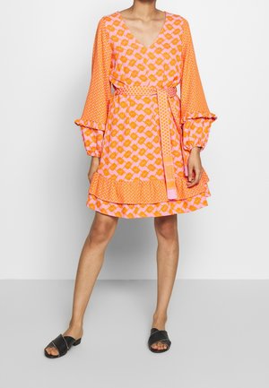 Vrouw draagt een roze en oranje patroon V-hals jurk met uitlopende mouwen, een taillekoord, gelaagde zoom en zwarte instapsandalen.