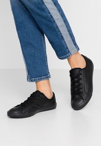 Personne portant des baskets basses noires et un jean en denim bleu avec une bande bleu clair, debout sur un sol blanc.