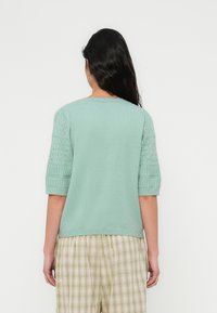 Πλεκτό πουλόβερ σε μέντα με κοντά μανίκια με υφή, διαθέτοντας ριμπ τελείωμα. Η πίσω πλευρά έχει λείο ύφασμα με ελάχιστες λεπτομέρειες.