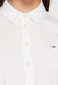 White button-up shirt featuring a classic collar, clear buttons, and a small embroidered logo in red, white, and blue on the left side.