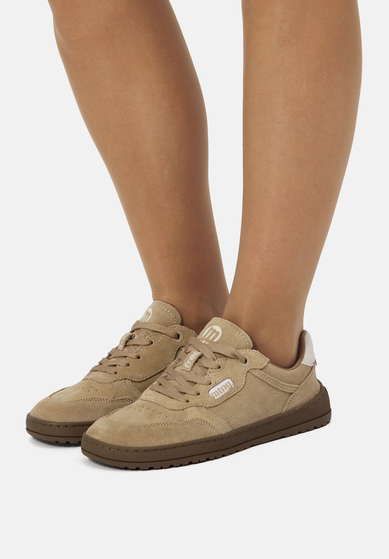 Zapatillas de gamuza de color marrón claro con cordones, detalles de logo en blanco, llevadas en los pies sobre un fondo blanco liso.
