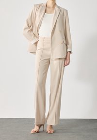 Beige op maat gemaakte pak met een dubbelzijdig colbert, wijde broekspijpen en textuurmanchetten. Gecombineerd met nude hoge hakken.