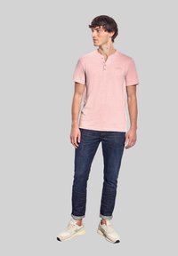 Růžové tričko Henley s krátkými rukávy a tříknopkovou légou, spárované s tmavými džínami a světle barevnými teniskami. Hladká textura.