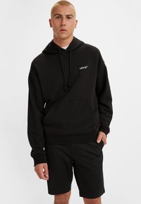 Svart hettegenser med frontlomme og snor, med en liten hvit Levi's-logo på brystet. Kombinert med svarte shorts.