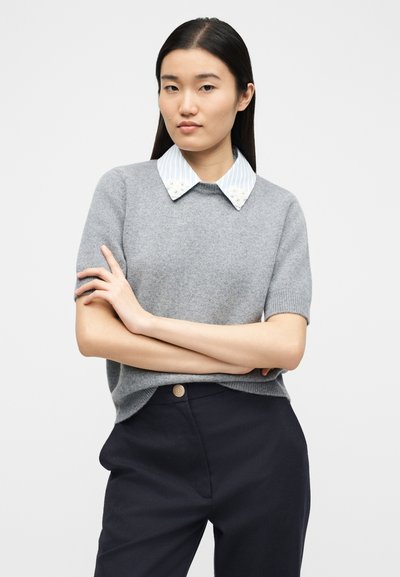 Femme aux longs cheveux bruns portant un pull gris à manches courtes par-dessus une chemise rayée avec un col floral, et un pantalon sombre, bras croisés.