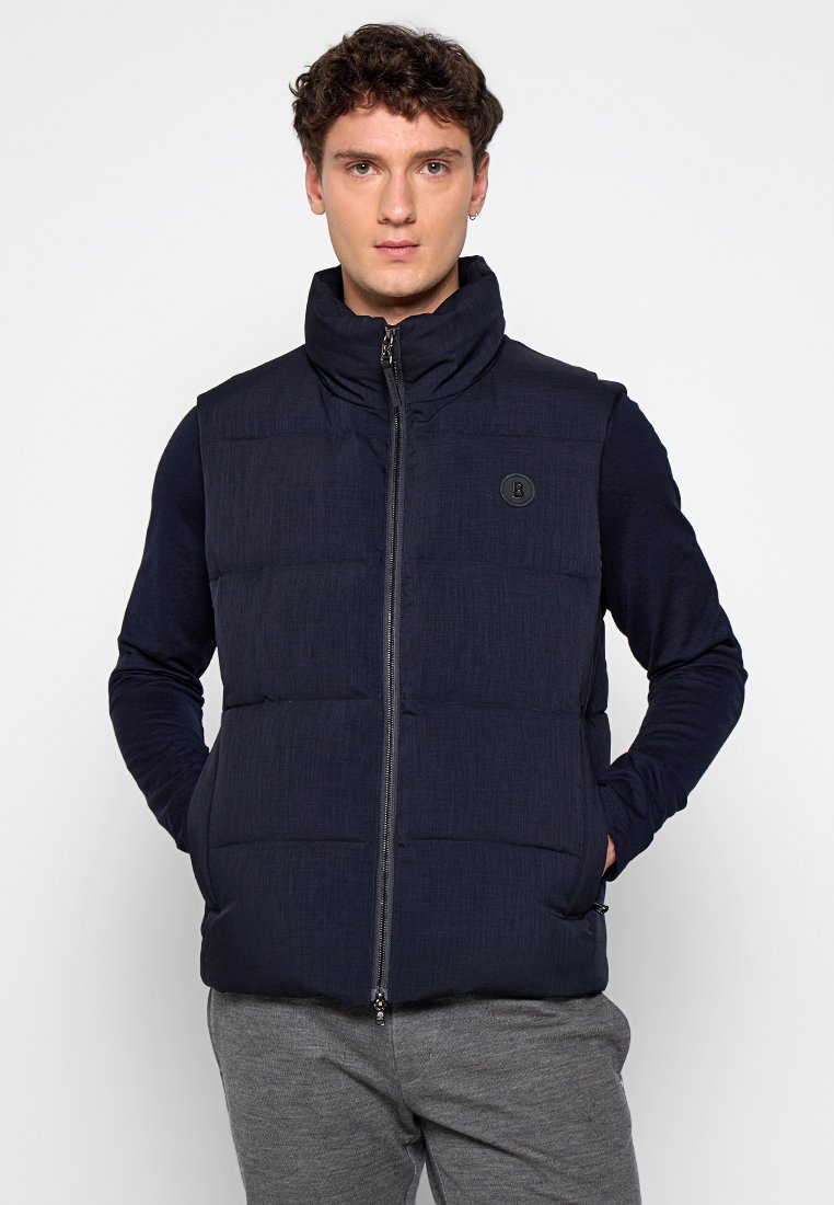 Dunkelblauer Puffer-Weste mit hohem Kragen, Reißverschluss vorne und strukturiertem Stoff. Mit Logo-Patch und zwei Seitentaschen, getragen über einem Langarmshirt.