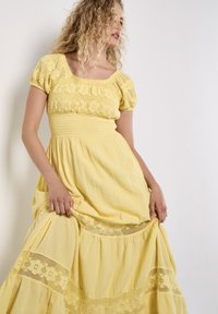 Een vrouw met krullend haar draagt een gele jurk met een smock-borsten, terwijl ze de zoom vasthoudt terwijl ze poseert tegen een lichte achtergrond.