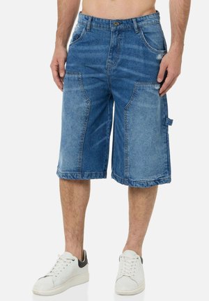 Uomo che indossa pantaloncini di jeans blu fino al ginocchio con grandi tasche frontali e scarpe da ginnastica bianche, in piedi su uno sfondo semplice.