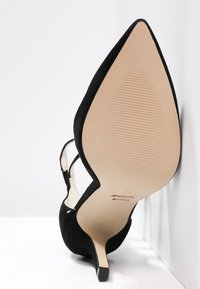 Černý semišový vysoký podpatek s špičatou špičkou a otevřenou stranou, s štíhlým stiletto podpatkem a hladkou béžovou podrážkou s drážkami.