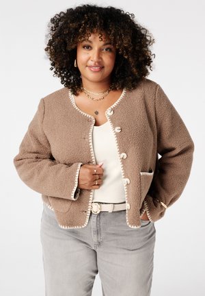 Femme aux cheveux bouclés portant une veste beige texturée à boutons sur un haut blanc, un pantalon gris et des bijoux en or, souriante avec les mains près des poches.