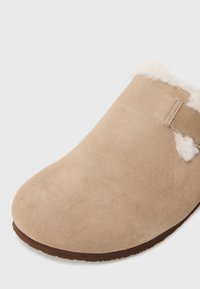Beige suede slipper with a rounded toe, featuring a soft fur trim at the opening and a durable brown rubber sole.