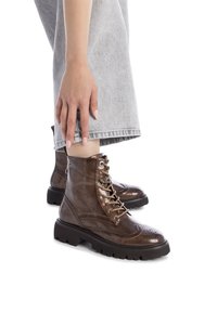 Botas de cuero marrón con cordones y detalles de brogue, con puntera redonda y suelas gruesas de goma negra, combinadas con pantalones de mezclilla gris claro.
