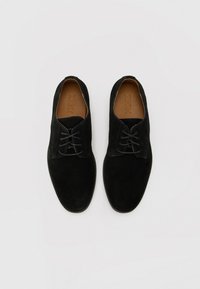 Sorte semskede snøresko med en glat tekstur, rund tå og minimalisk design. Har en brun sål med merkevarenavnet "ZIGN."