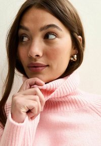 Roze geribbelde coltrui met een zachte textuur, voorzien van een hoge kraag. Model trekt voorzichtig met één hand aan de kraag.