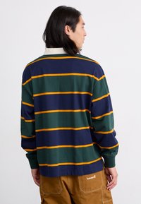 Long-sleeve polo shirt with green, navy, and yellow stripes, featuring a white collar and soft cotton material. Timberland logo on back.