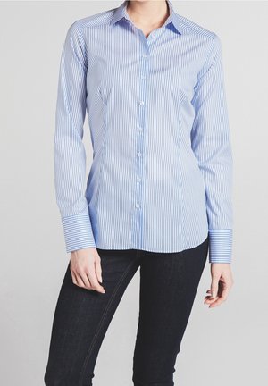 Frau trägt eine eng anliegende blau-weiß vertikal gestreifte Bluse mit langen Ärmeln und dunkle, schmale Jeans.