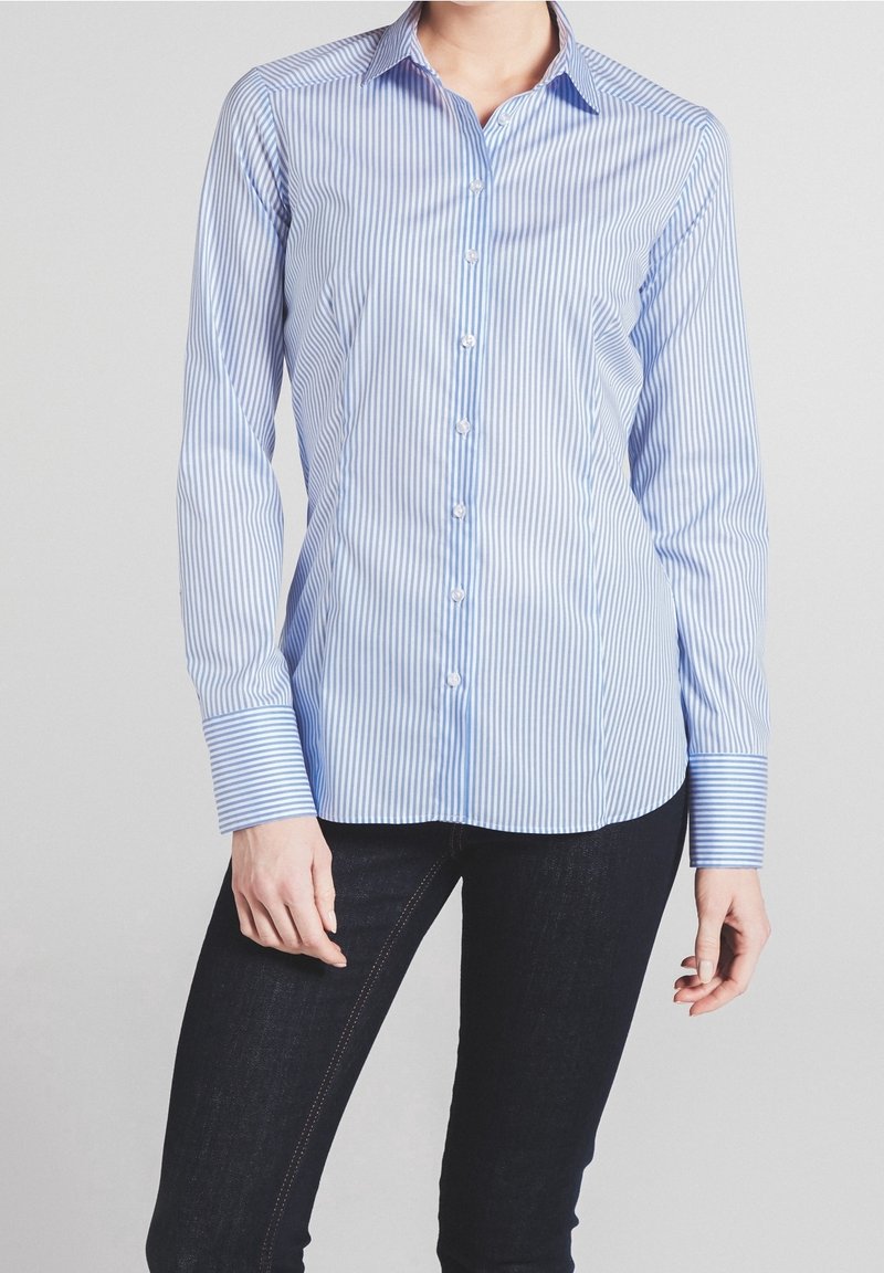 Femme portant une chemise cintrée rayée verticalement bleu et blanc à boutons, manches longues, et un jean slim foncé.
