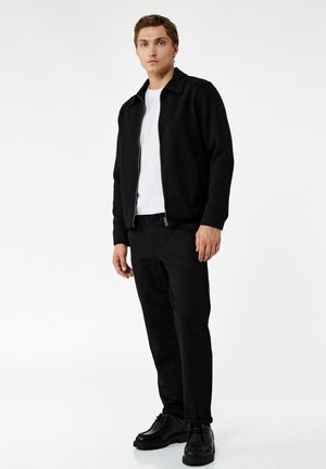 Jonge man die een zwart jack met rits, een wit T-shirt, zwarte broek en zwarte schoenen draagt, staand tegen een effen witte achtergrond.