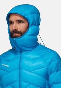 Giacca puffer blu con cappuccio, caratterizzata da un design trapuntato, colletto alto e chiusura con zip. Marchio visibile sul lato sinistro del petto.