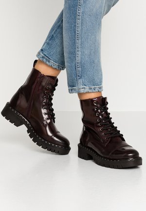 Botas de combate brillantes de color burdeos oscuro con cordones y cremallera lateral, usadas con vaqueros azul claro remangados sobre un suelo blanco brillante.