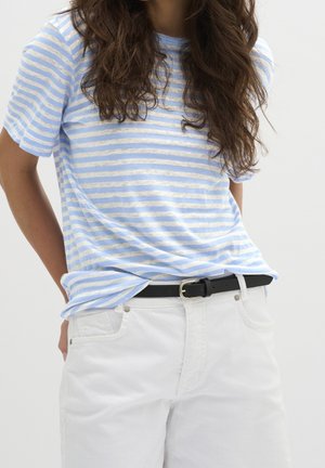 Femme portant une chemise à manches courtes rayée bleu clair et blanche rentrée dans un pantalon blanc avec une ceinture noire.