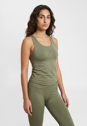 Kvinde med bølget mørkt hår iført en tætsiddende olivengrøn ærmeløs top og matchende leggings, stående mod en ensfarvet lys grå baggrund.