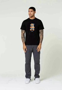 T-shirt de algodão preta com uma gráfica colorida de um urso na frente. O modelo combina com calças cargo cinzas e ténis brancos.