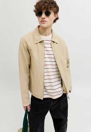 Mladić sa zaobljenim sunčanim naočalama, bež jaknom preko bijele košulje s crvenim prugama, crne hlače, drži zelenu torbu na jednobojnoj pozadini.