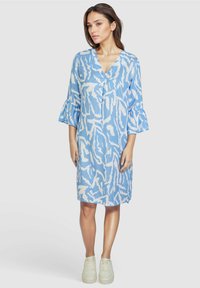 Blaues Kleid mit V-Ausschnitt, dreiviertel langen Glockenärmeln und einem weißen abstrakten Blattmuster. Hergestellt aus leichtem Stoff. Getragen mit weißen Sneakers.