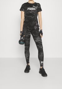Conjunto deportivo de PUMA con patrón de mármol negro y gris, que incluye parte superior y leggings con logo, combinado con zapatos negros y guantes de agarre, sosteniendo una kettlebell.