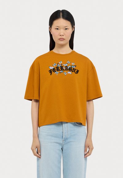 Πορτοκαλί βαμβακερό T-shirt με στρογγυλή λαιμόκοψη και κοντά μανίκια, διακοσμημένο με ένα λουλουδάτο γραφικό και το κείμενο "FERRAGNI" σε μαύρο χρώμα. Φοριέται με ανοιχτού χρώματος μπλε τζιν.