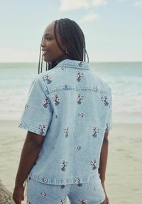 Cămașă din denim bleu deschis, cu mâneci scurte, având broderie florală în roz și navy, expusă pe un fundal de plajă.