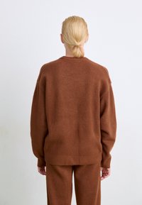 Brun strikket genser med løs passform, rund hals og ribbestrikkede mansjetter, kombinert med matchende brun underdel, vist bakfra.