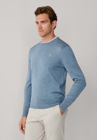 Lichtblauwe trui, ronde hals, geribbelde manchetten en zoom, gemaakt van zacht materiaal. Voorzien van een klein logo op de borst, gecombineerd met beige broek.