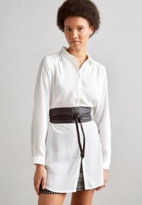 Witte overhemd met lange mouwen, voorzien van een zwarte gevlochten leren riem. Rok heeft een gestructureerd patroon, knie-lengte, gladde stof.