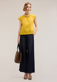 Blusa gialla a maniche corte con dettagli a pieghe, abbinata a pantaloni a gamba larga blu navy. Gli accessori includono una borsa marrone testurizzata e scarpe con plateau a fantasia.
