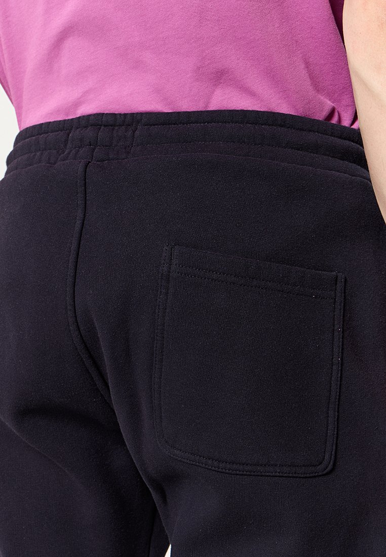 Pantaloni della tuta neri realizzati in tessuto morbido, con vita elastica e una singola tasca posteriore. La texture appare liscia e accogliente.