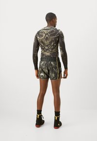 Long-sleeve black compression top with a detailed gold skull design; black shorts with camouflage pattern and logo; black and yellow athletic shoes.
