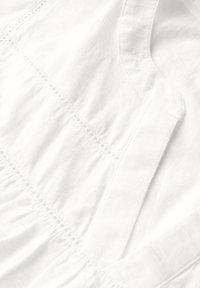Tejido blanco con acabado texturizado, que presenta costuras delicadas y volantes. El diseño incluye un escote redondo y detalles estructurados en el cuerpo.