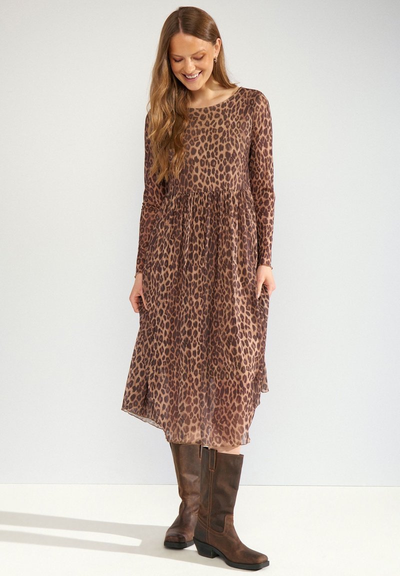 Femme portant une robe midi marron à manches longues imprimée léopard, associée à des bottes hautes marron foncé, debout et regardant vers le bas avec un sourire.