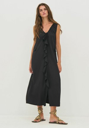 Kvinde står foran en ensfarvet baggrund iført en sort midi-kjole uden ærmer med frontflæse og bindedetalje, kombineret med gyldne sandaler med nitter.